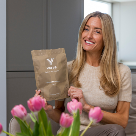 A smiling woman sitting indoors holding a pouch of Verve Honest Mushrooms Mix 21, with pink tulips in the foreground, conveying a positive and uplifting wellness moment.