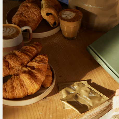 Two lattes, croissants, pain au chocolat, verve mushroom drink sachets, and a green book on a wooden table in natural light.