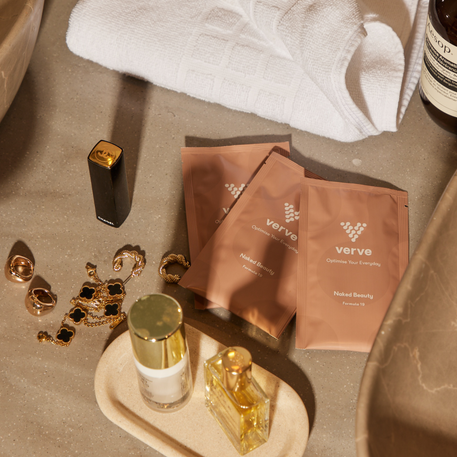 Three Verve sachets, jewellery, perfume bottles, a Chanel lipstick and a folded white towel on a bathroom counter.