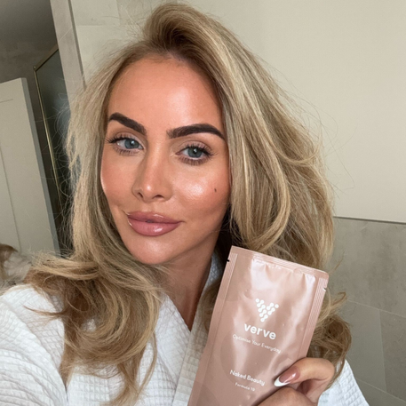 A woman in a white robe taking a bathroom selfie while holding a sachet of Verve Naked Beauty Collagen Formula 19, showcasing glowing skin and healthy beauty routine.