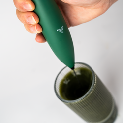 Hand holding a green electric whisk, mixing a dark green drink in a ribbed glass.