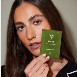 A woman holding a green Verve “Transparent Greens” sachet close to her face, standing against a neutral background.