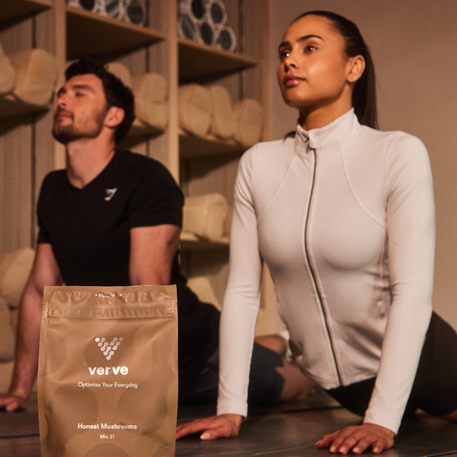 A pouch of Verve Honest Mushrooms Mix 21 placed in the foreground, with two people in yoga poses behind it, promoting focus, energy, and wellness in a calming studio setting.