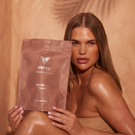 A woman with glowing skin holding a pouch of Verve Naked Beauty Collagen Formula 19, seated in a warm-toned setting, showcasing radiant wellness and skincare benefits.